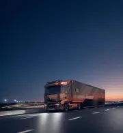A Renault Trucks T High tractor unit pulling a motor racing semi-trailer is travelling along a motorway at dusk, near a floodlit circuit.